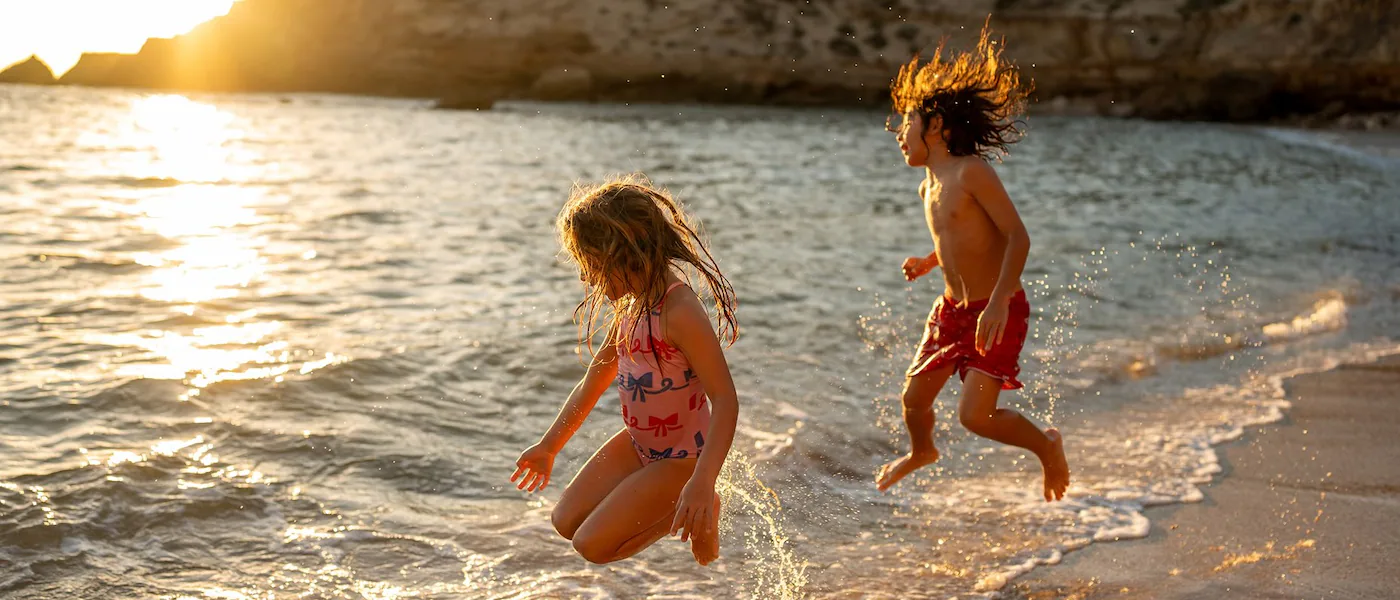 Children jumping over the tide as it rolls into the beach, with rocky coastline in the background.