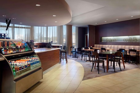 A modern hotel lobby with a curved reception desk, a beverage dispenser, and a seating area with tables and chairs.
