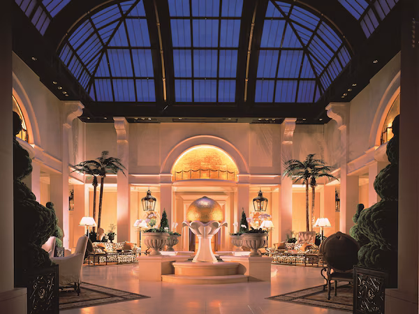A spacious lobby with a high ceiling and a skylight, featuring a central fountain, elegant seating, and palm trees.