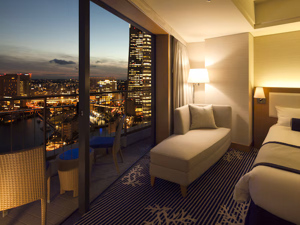 A hotel room with a city view, a balcony with wicker furniture, and a bed with a headboard.