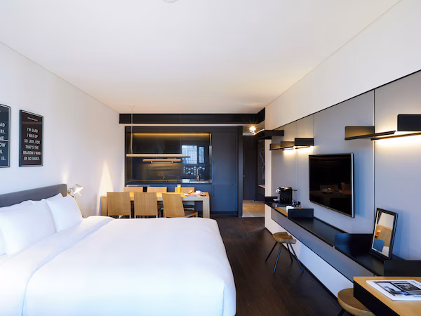 A modern hotel room with a large bed, a dining area, a kitchenette, and a TV mounted on the wall.