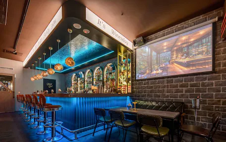 A bar area with a blue-lit bar counter, multiple stools, and a projection screen displaying a bar scene.