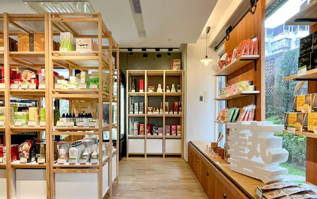 A store interior with wooden shelves stocked with various products, a counter with items, and a view of greenery outside.