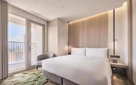 A modern hotel room with a large bed, a seating area, and a balcony with a city view.