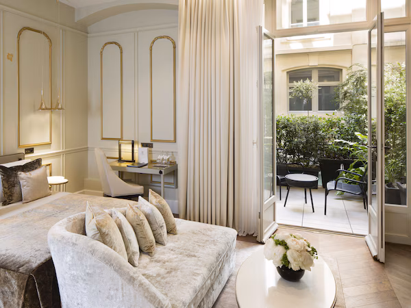 A luxurious bedroom with a large bed, a sofa, a desk, and a view of an outdoor patio with plants and furniture.