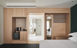 A modern hotel room with a wooden wardrobe, a small desk with a kettle, a bed, and a mirror.