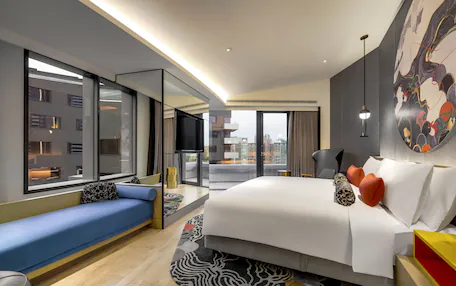 A modern hotel room with a large bed, a seating area, a large window, and a city view.