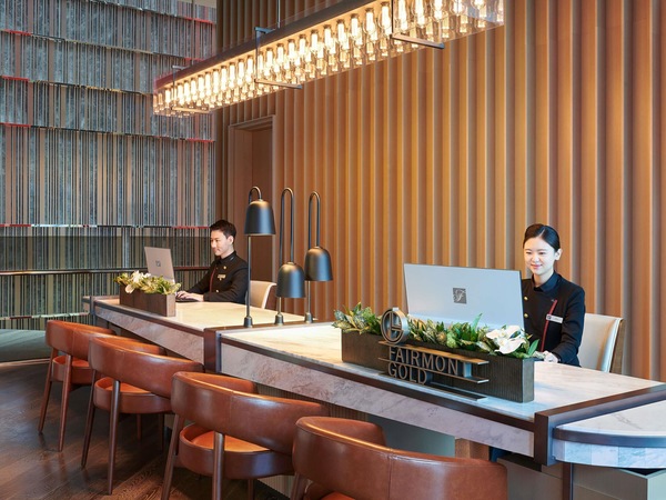A modern hotel reception area with a wooden panel wall, two receptionists at desks, and a large, illuminated light fixture above.