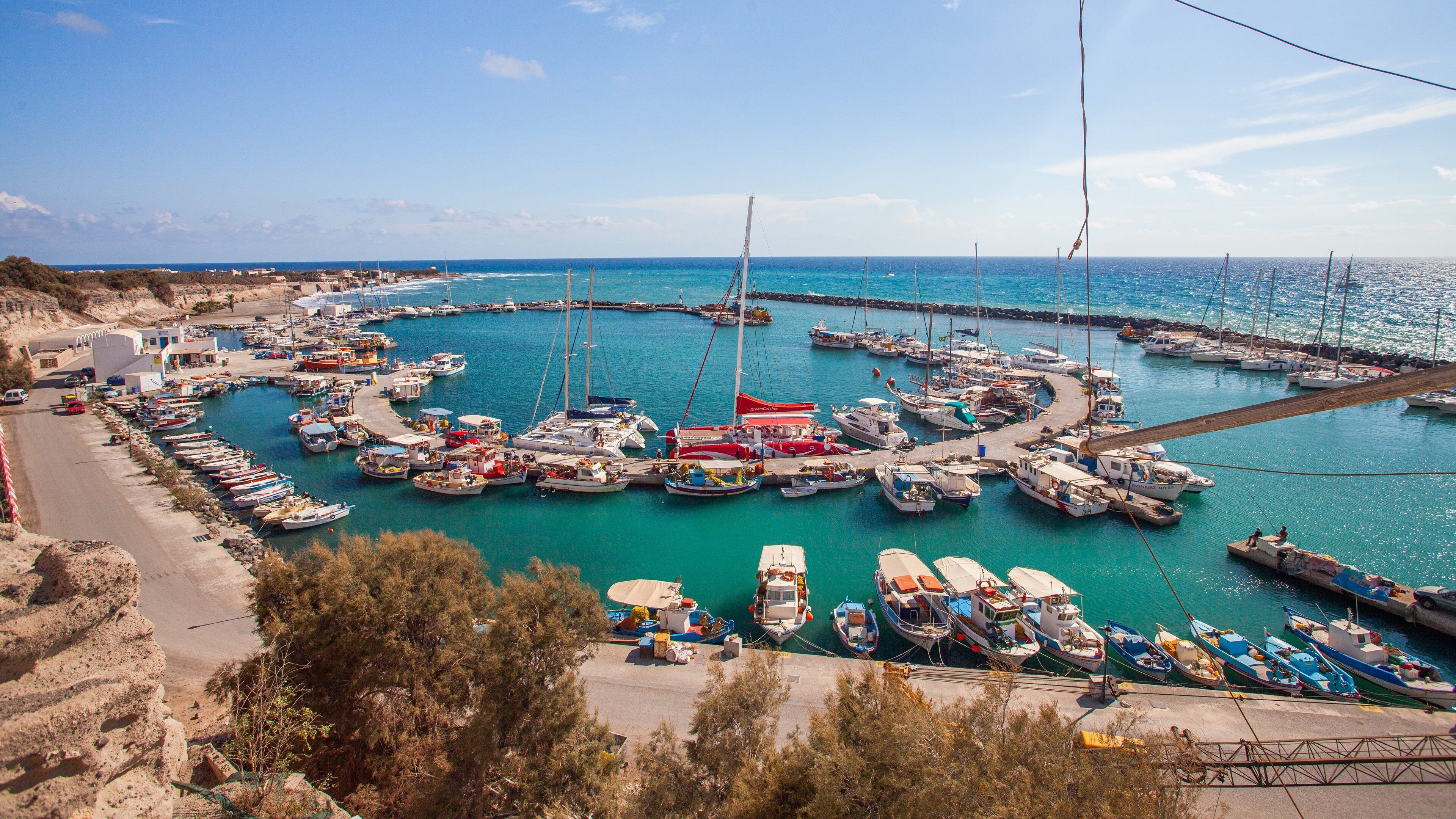 A marina with numerous boats docked in a circular arrangement, surrounded by a coastal landscape and a clear blue sky.