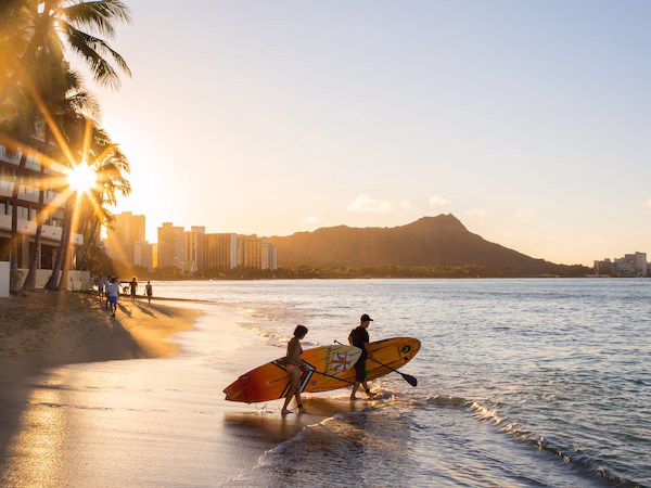 A beach with palm trees, a sunset, and surfers carrying surfboards.